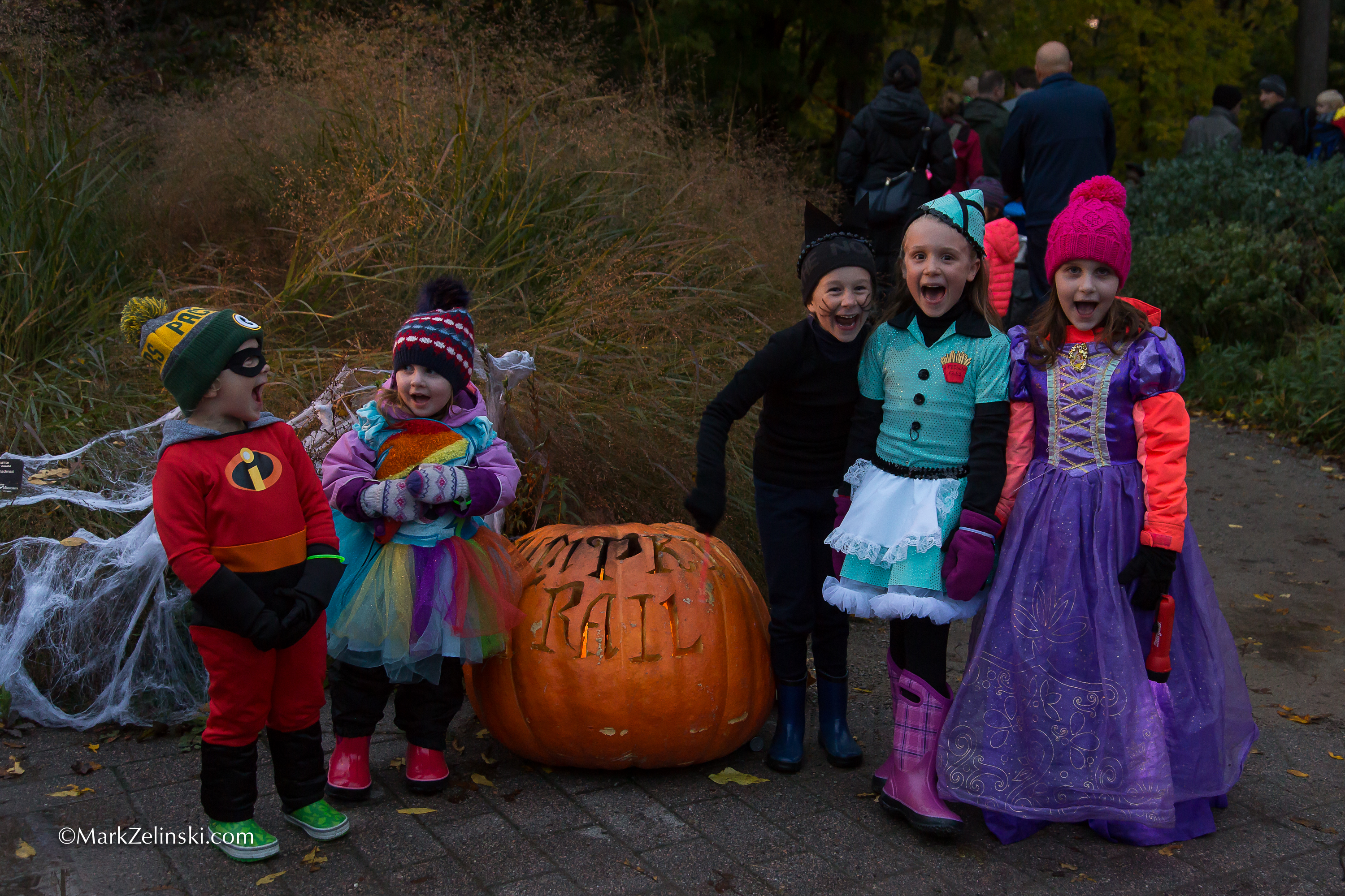 children in costume standing alongside a giant jack-o-lantern carved with the words "Pumpkin Trail"